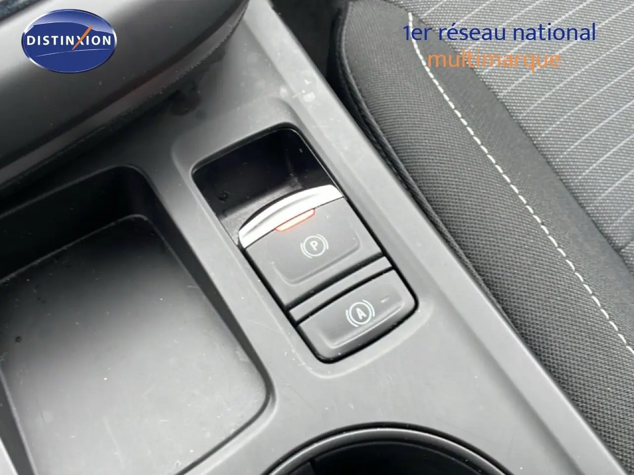 Gros plan sur la console centrale montrant les boutons de frein à main électrique et de démarrage-arrêt automatique, siège tissu noir visible.