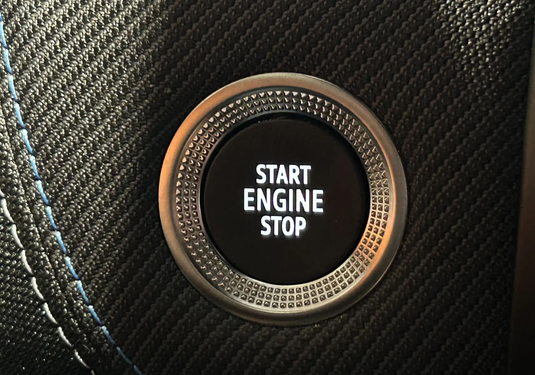Gros plan sur le bouton Start Engine Stop entouré d’un cerclage métallique et d’un habillage noir avec surpiqûres bleues.