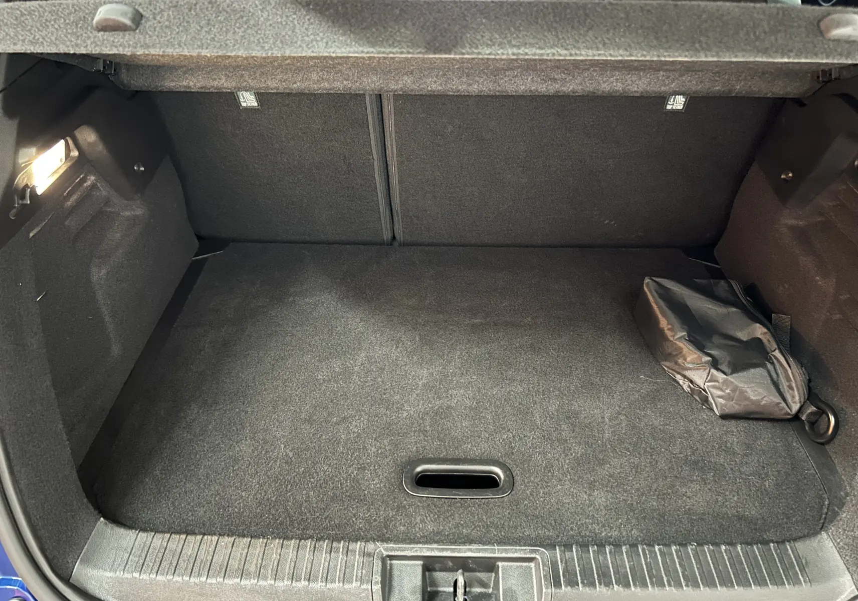 Coffre ouvert vu de face d’un Renault Captur bleu Iron, avec tapis noir et sac de rangement sur le côté droit.