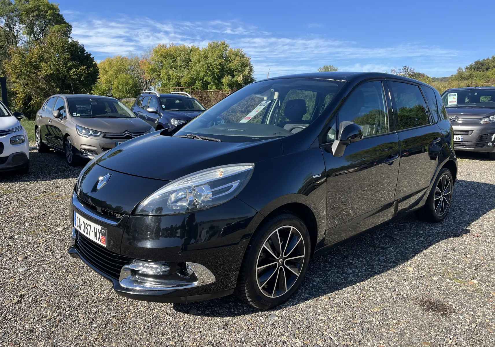Renault Scenic III noir en 3/4 avant droit, avec jantes alliage et calandre chromée brillante sous un ciel dégagé.