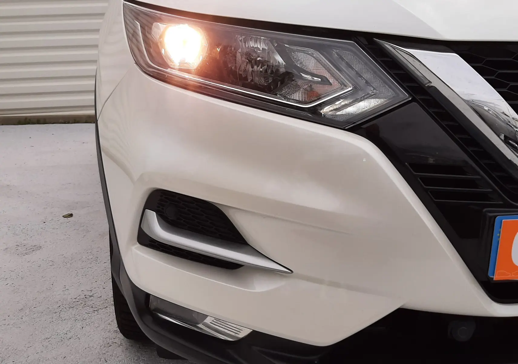 Gros plan sur l'avant gauche d'un Nissan Qashqai blanc 2020 avec phare allumé et calandre noire brillante.