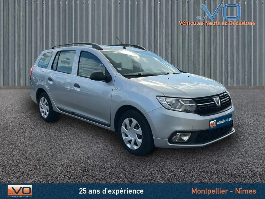 Vue 3/4 avant droite d'une Dacia Logan MCV gris clair avec barres de toit et jantes 15 pouces, stationnée sur sol bitumé.
