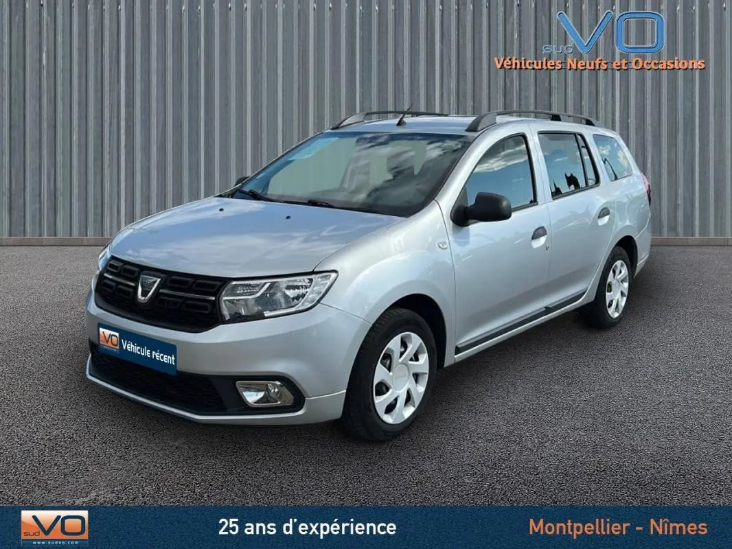 Dacia Logan MCV gris clair vue 3/4 avant droit avec barres de toit et jantes 15 pouces sur parking.