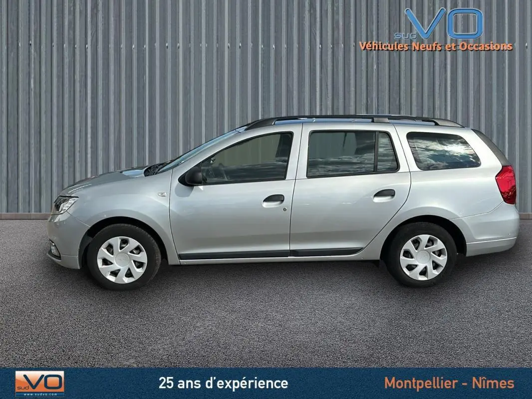 Profil côté gauche d'une Dacia Logan MCV gris clair avec barres de toit, garée devant un mur métallique.
