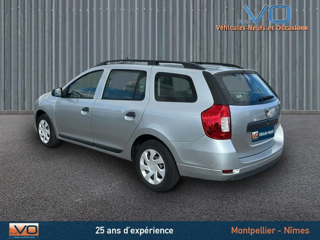 Vue 3/4 arrière droite d'une Dacia Logan MCV gris clair avec barres de toit et jantes 15 pouces en environnement urbain.