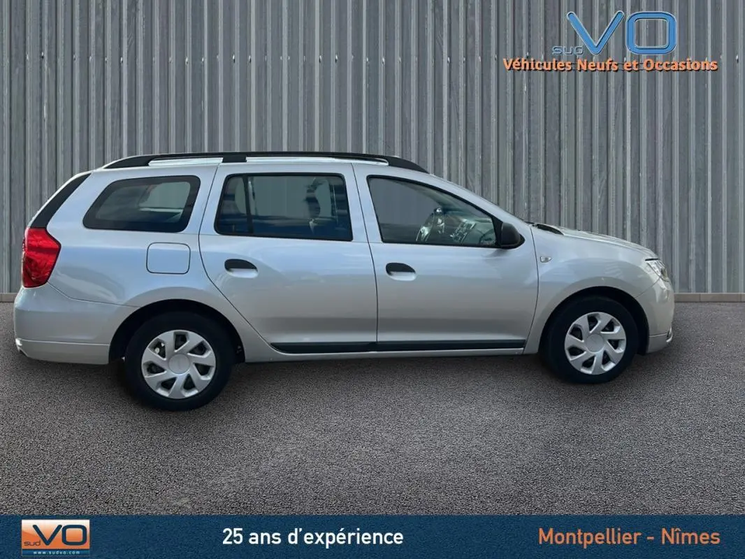 Profil côté droit d'une Dacia Logan MCV gris clair avec barres de toit, garée devant un mur métallique.