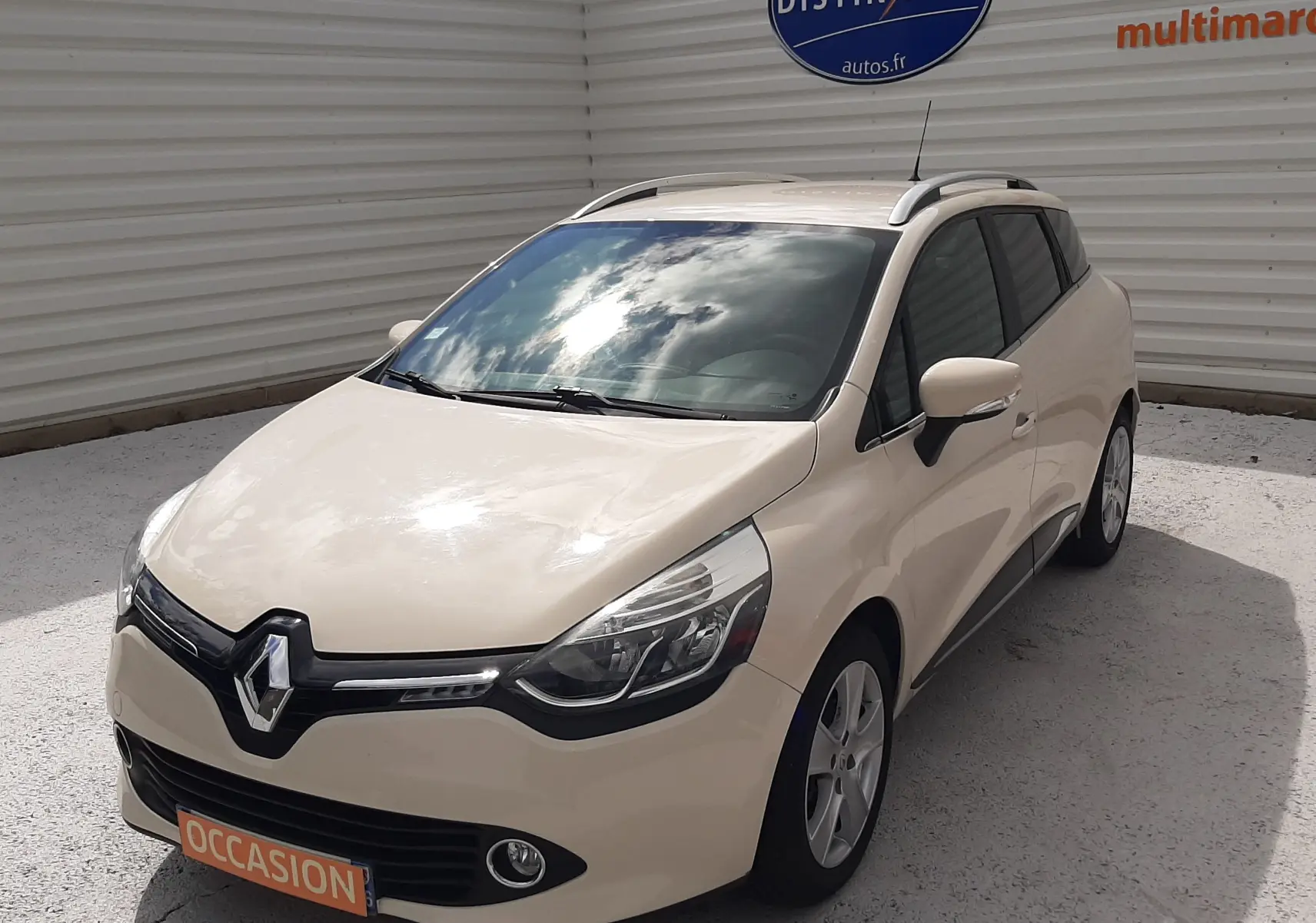 Renault Clio IV Estate beige vue 3/4 avant droit, avec toit ouvrant et barres de toit visibles.