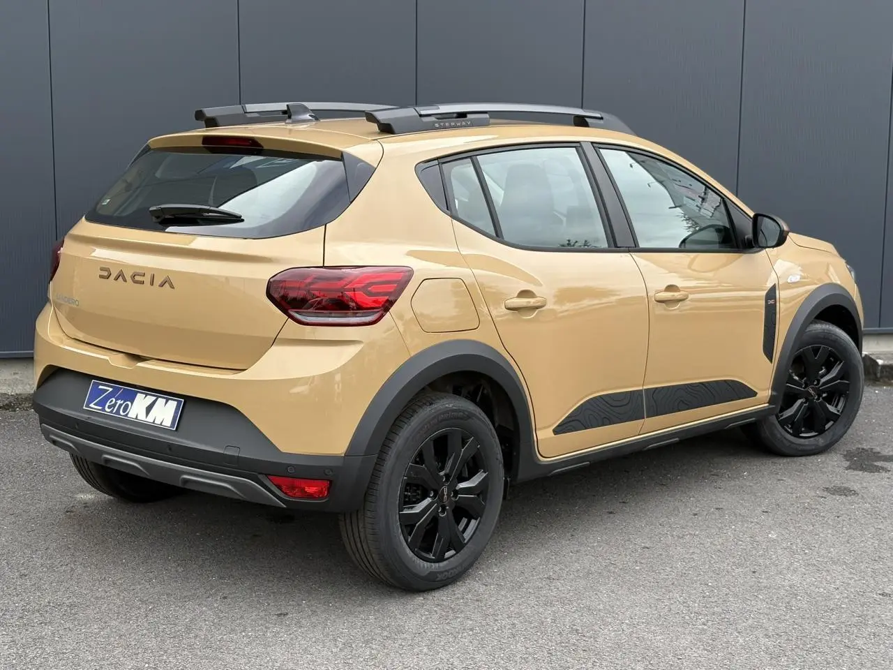 Vue 3/4 arrière droite d'une Dacia Sandero Stepway beige Safari avec jantes noires et barres de toit grises.