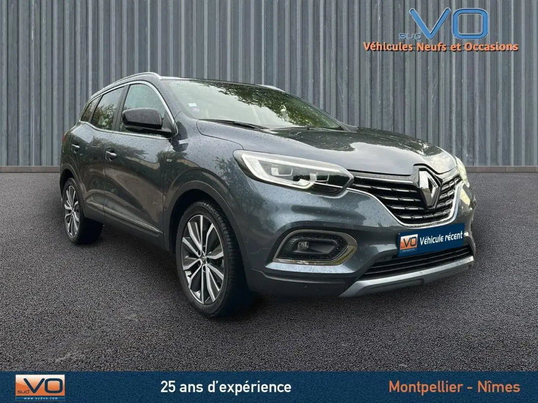 Renault Kadjar gris 3/4 avant droit avec jantes alliage 18 pouces et barres de toit visibles sur sol asphalté.