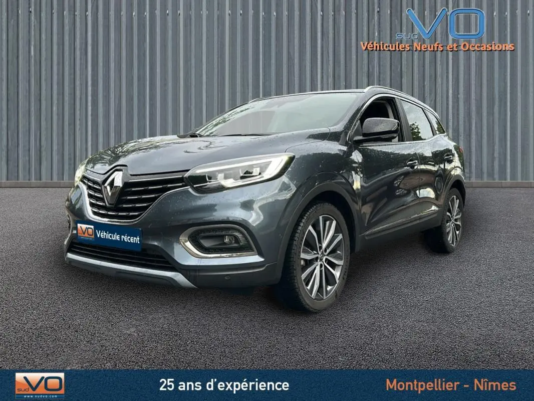 Renault Kadjar gris 3/4 avant droit avec jantes alliage 18 pouces et barres de toit visibles sur fond urbain.
