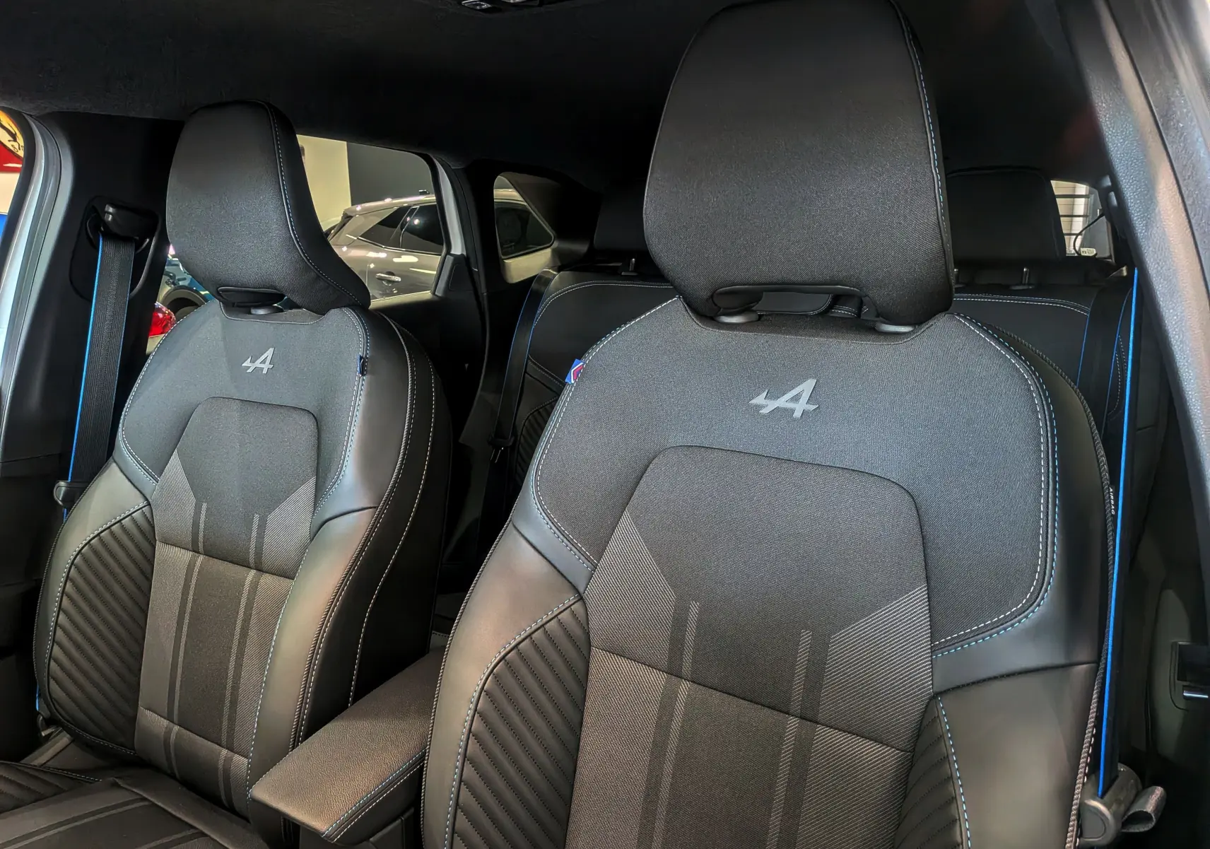 Gros plan sur les sièges avant noirs avec surpiqûres bleues et logo Alpine dans l’habitacle du Renault Symbioz blanc nacré.
