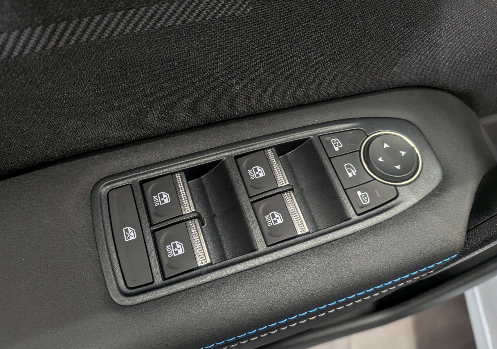 Gros plan sur les commandes de lève-vitres et rétroviseurs sur la porte côté gauche, intérieur noir avec surpiqûres bleues.