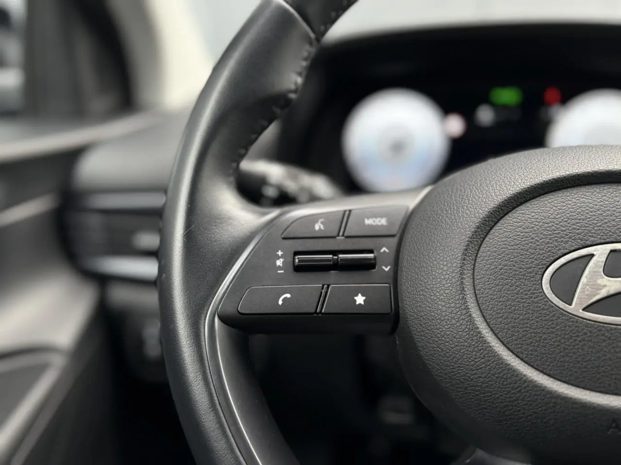 Gros plan sur le volant cuir noir de la Hyundai i20 2025 avec commandes intégrées, tableau de bord numérique flou en arrière-plan.