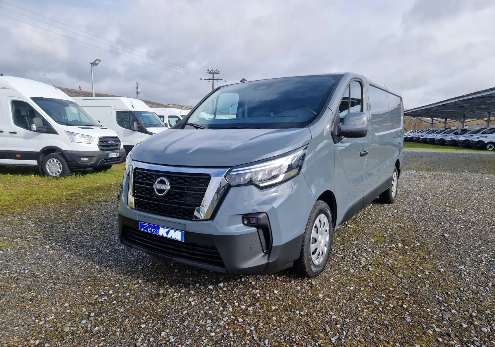 Nissan Primastar gris urbain en vue 3/4 avant droit sur un terrain gravillonné avec plusieurs fourgons blancs en arrière-plan.