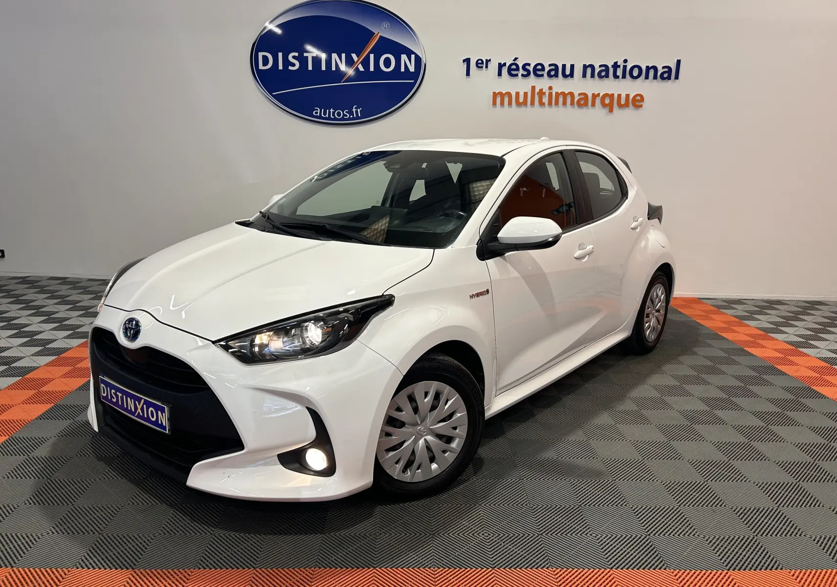 Toyota Yaris hybride blanche vue 3/4 avant droit en intérieur avec phares et antibrouillard allumés.