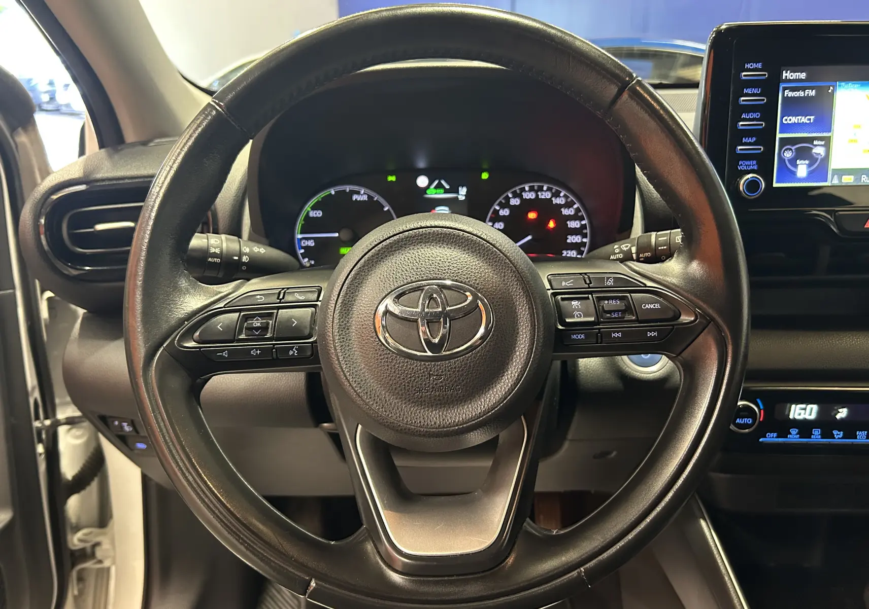 Vue centrée sur le volant noir de la Toyota Yaris 116h blanche, avec tableau de bord numérique et écran tactile visible.