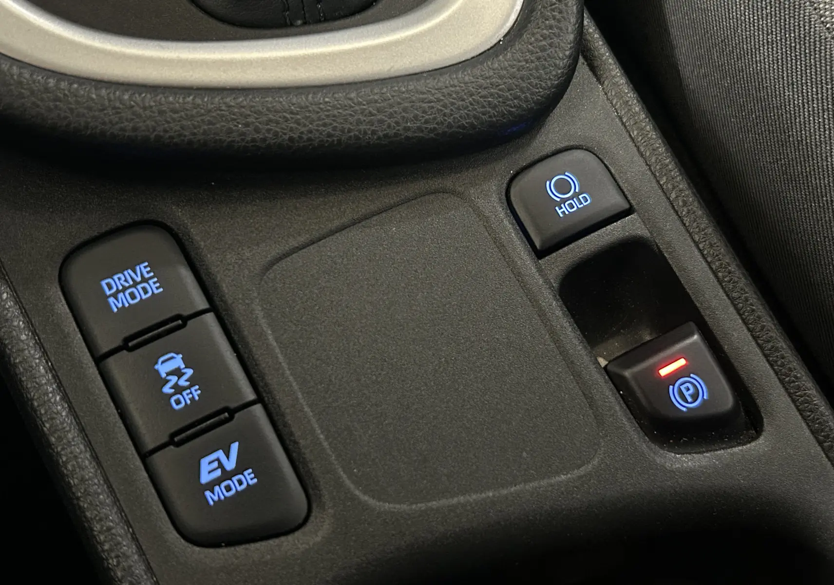 Gros plan sur la console centrale noire de la Toyota Yaris 116h, avec boutons lumineux Drive Mode, Hold et frein à main électrique.