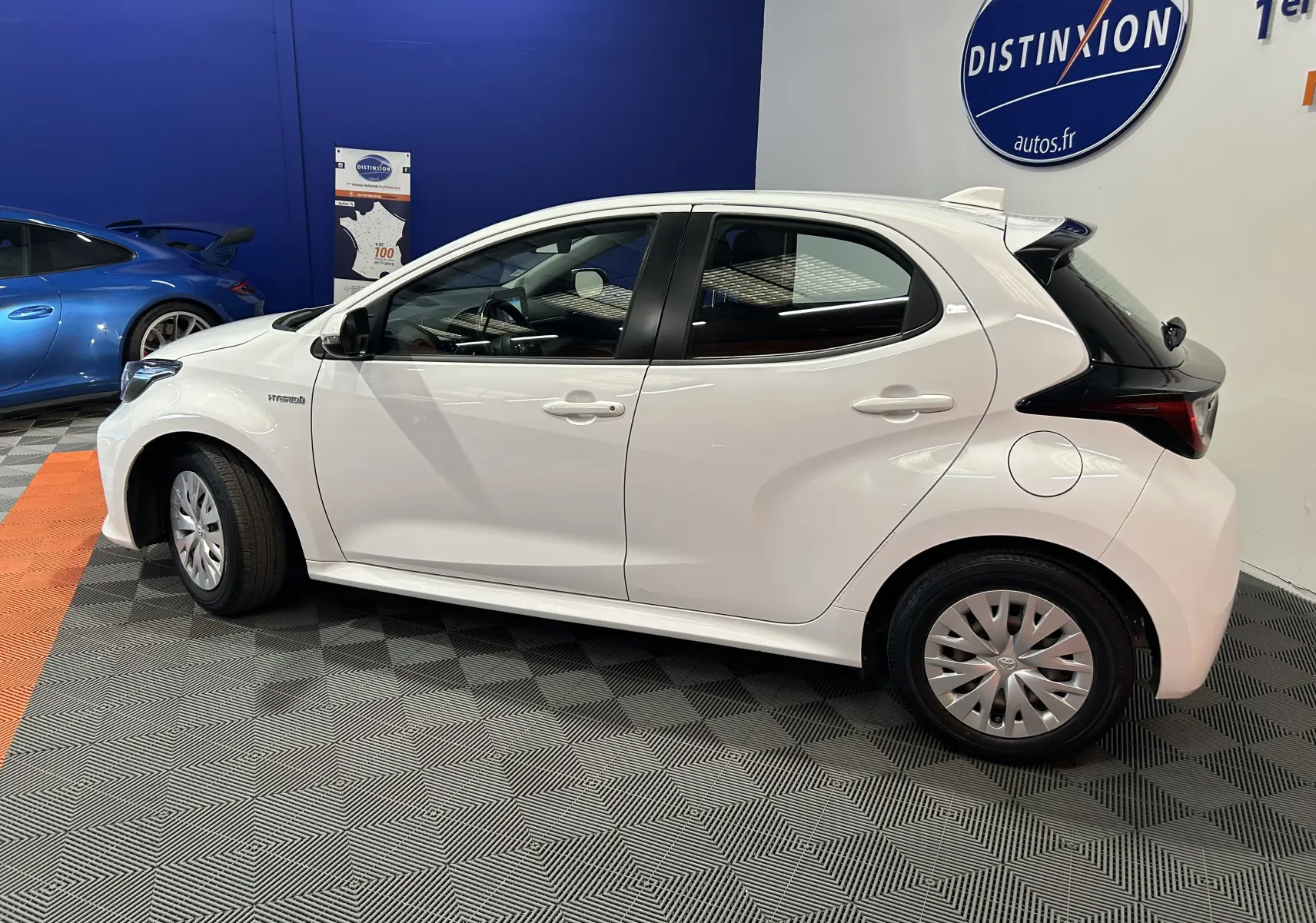 Profil côté gauche d'une Toyota Yaris hybride blanche 5 portes, en intérieur showroom avec logo Distinxion en arrière-plan.