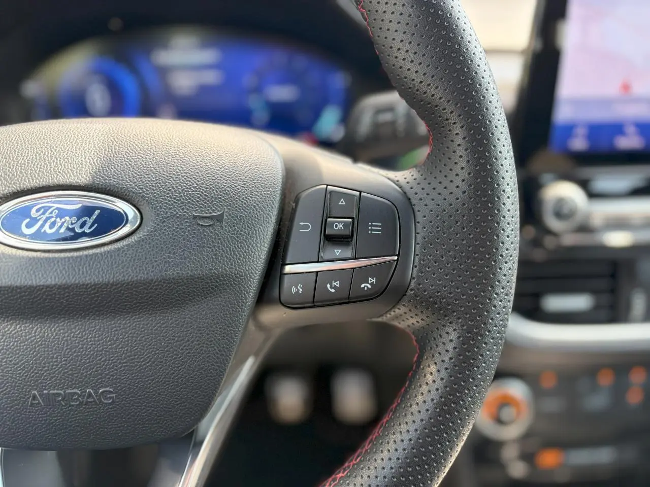 Gros plan sur le volant perforé avec surpiqûres rouges du Ford Puma ST Line, tableau de bord numérique et écran GPS en arrière-plan flou.