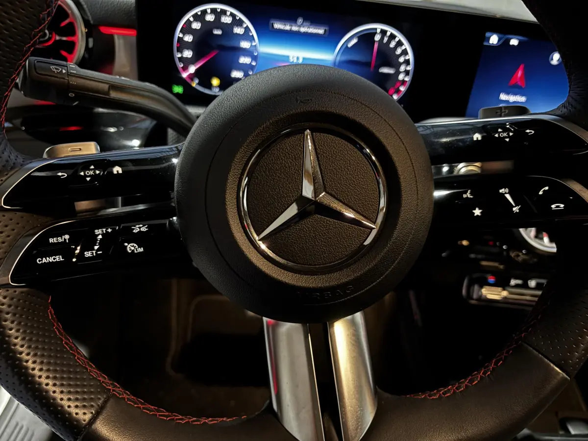 Gros plan sur le volant cuir noir avec surpiqûres rouges de la Mercedes CLA 250 e AMG Line, tableau de bord digital en arrière-plan.