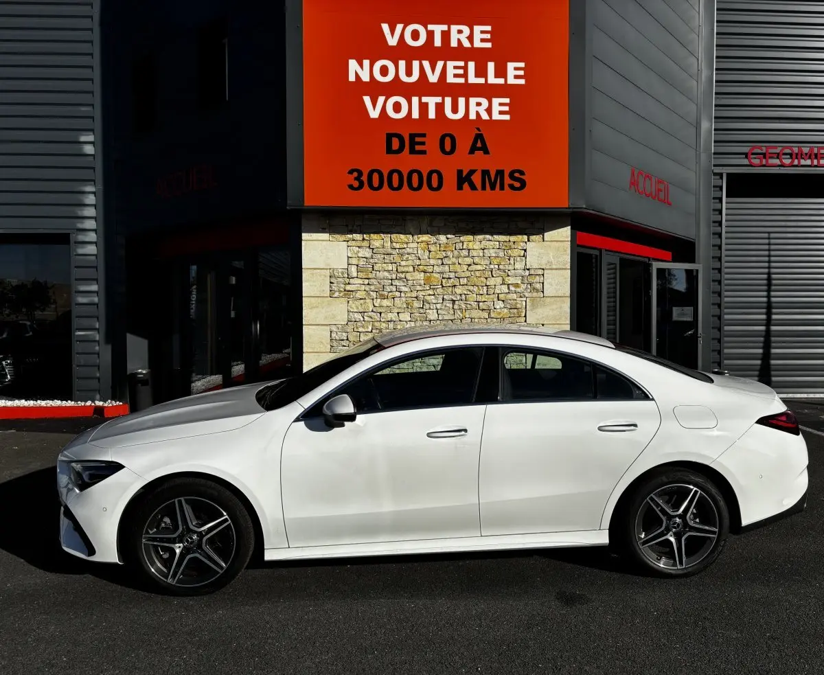 Vue de profil côté gauche d'une Mercedes CLA 250 e AMG Line blanche stationnée devant un bâtiment avec panneau publicitaire rouge.