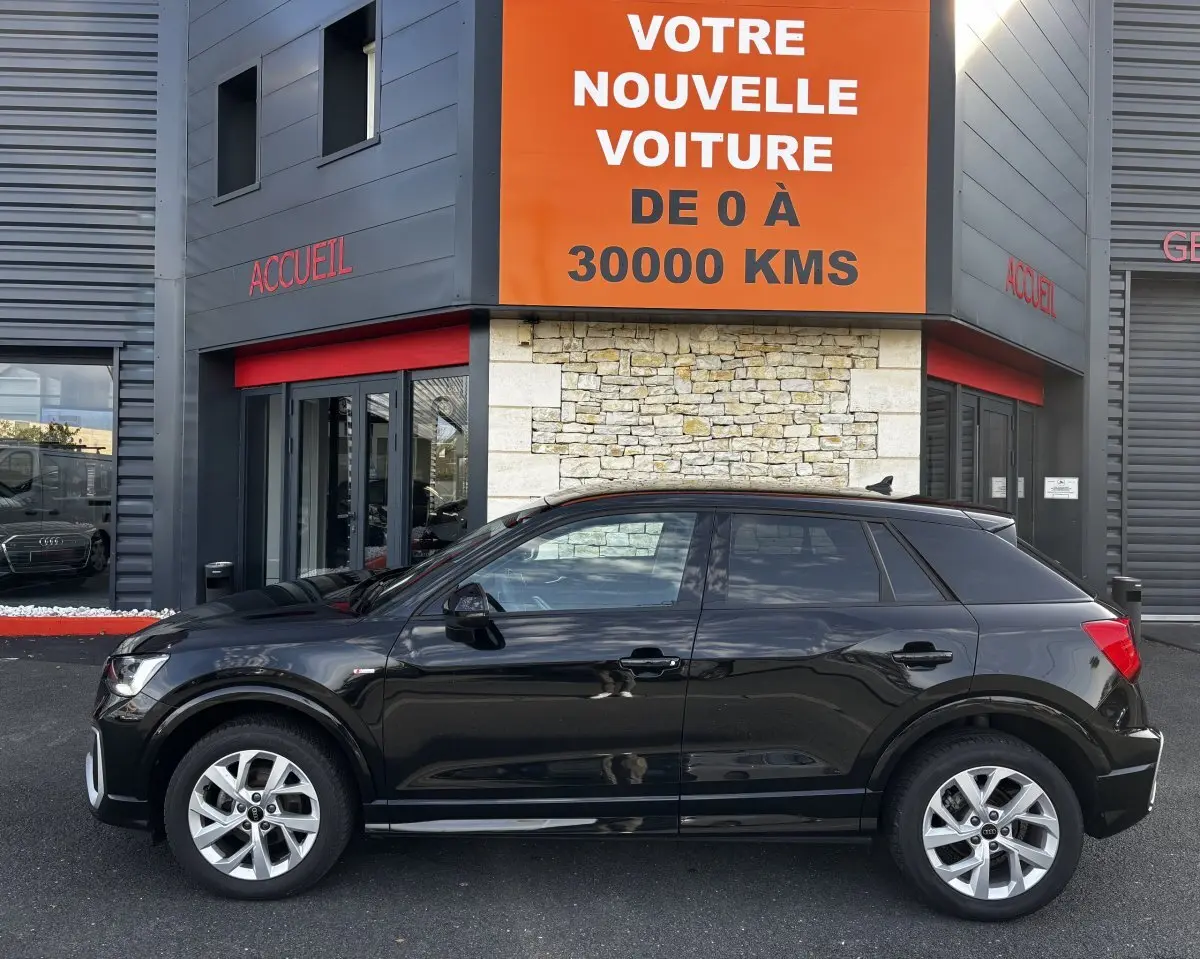 Audi Q2 noir vue de profil côté gauche, garée devant un bâtiment avec panneau orange et gris.