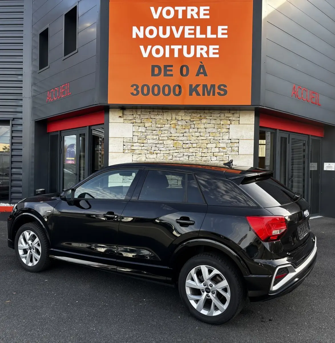 Audi Q2 35 TFSI S Line noir vue 3/4 arrière droit, stationnée devant un bâtiment avec panneau orange.