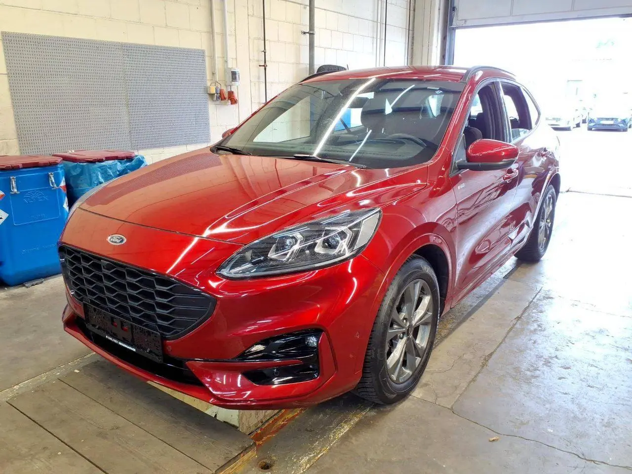 Ford Kuga 2024 rouge Lucifer en 3/4 avant droit, avec calandre noire et jantes alliage visibles en intérieur d'atelier.