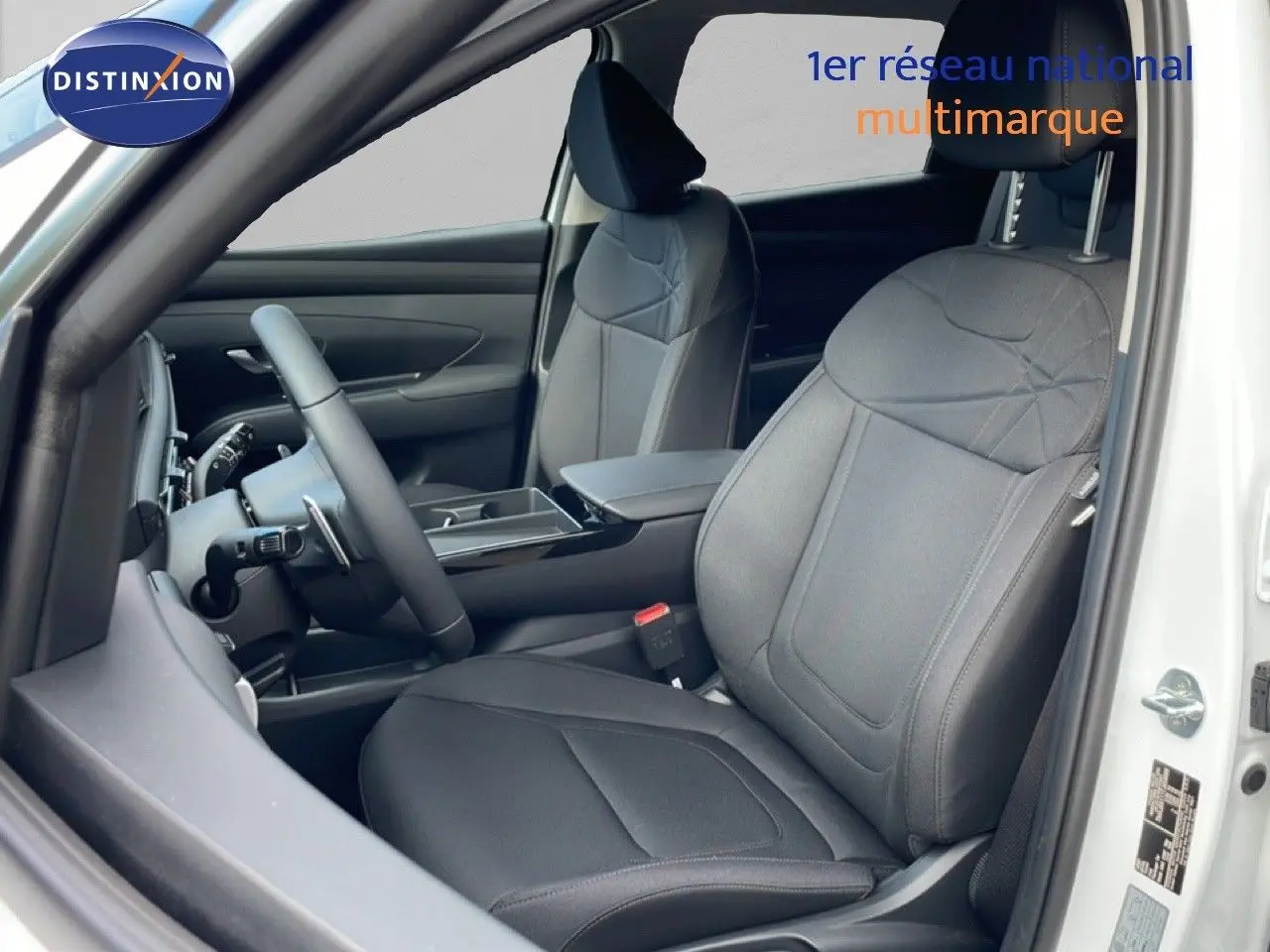 Vue intérieure côté conducteur du Hyundai Tucson 2026 blanc Atlas, sièges tissu noir avec détails couture et volant multifonction.