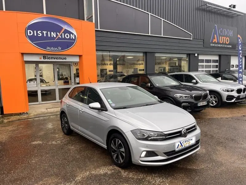 Volkswagen Polo gris 3/4 avant droit, avec jantes alu et calandre chromée, stationnée devant un concessionnaire.