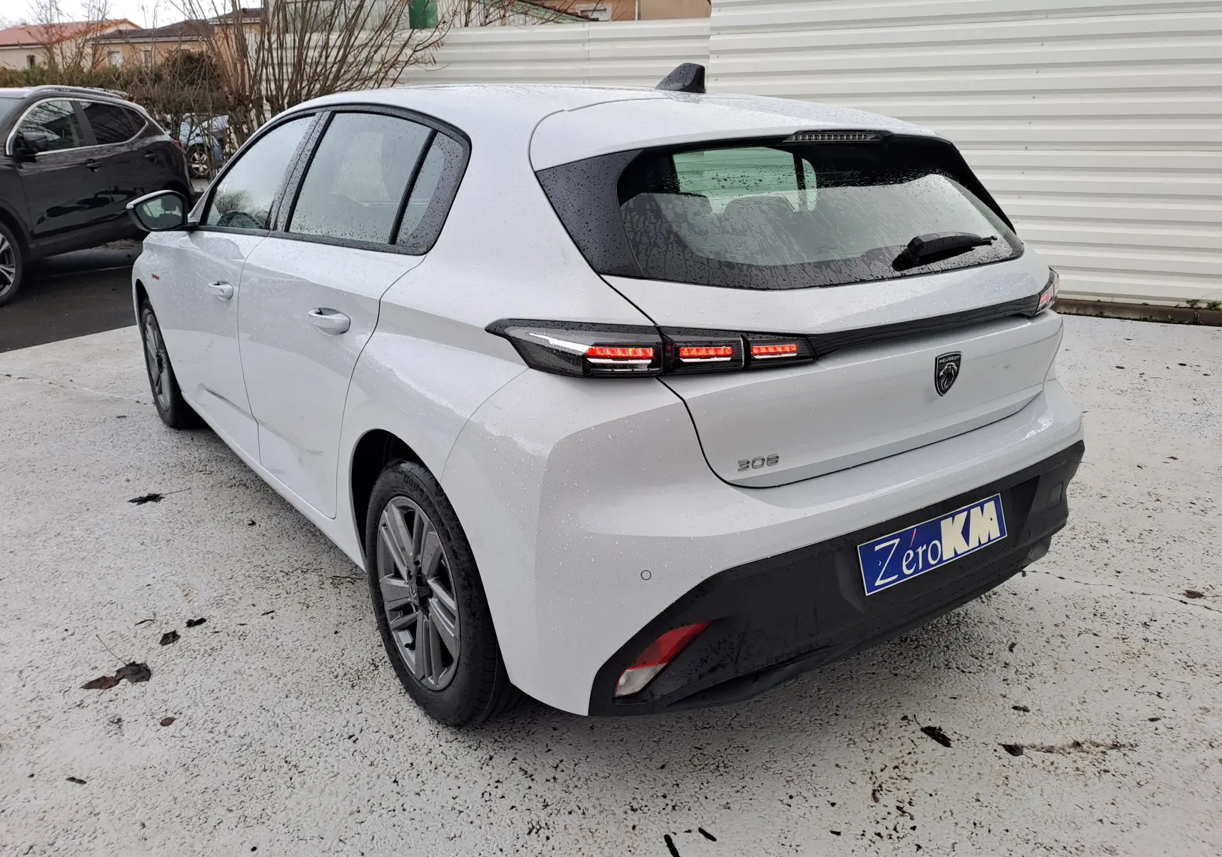 Vue 3/4 arrière droite d'une Peugeot 308 blanche Okenite avec feux arrière allumés et plaque d'immatriculation Zero KM.