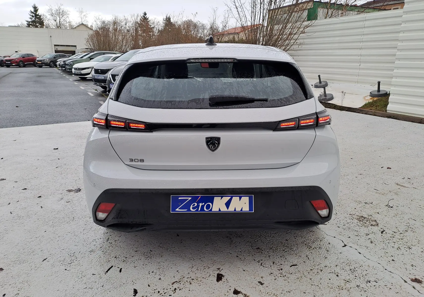 Vue arrière d'une Peugeot 308 blanche Okenite 2025 avec feux arrière LED allumés sur parking extérieur.
