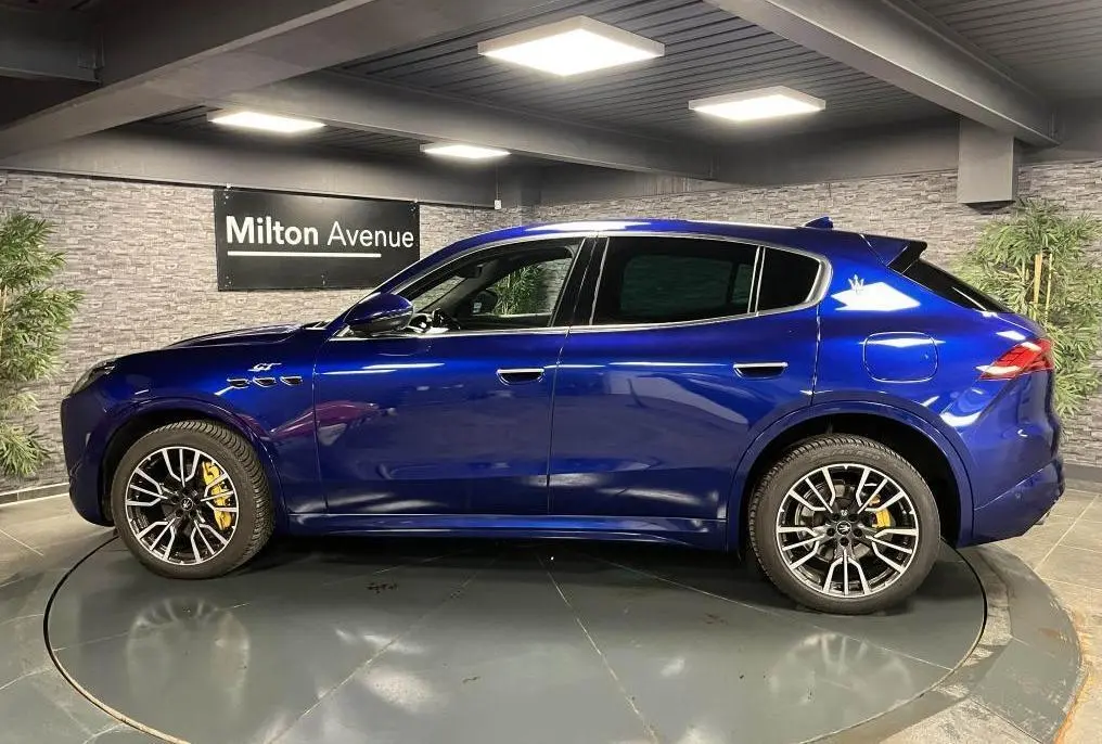 Profil droit du Maserati Grecale bleu avec jantes noires et étriers de freins jaunes visibles en intérieur showroom.