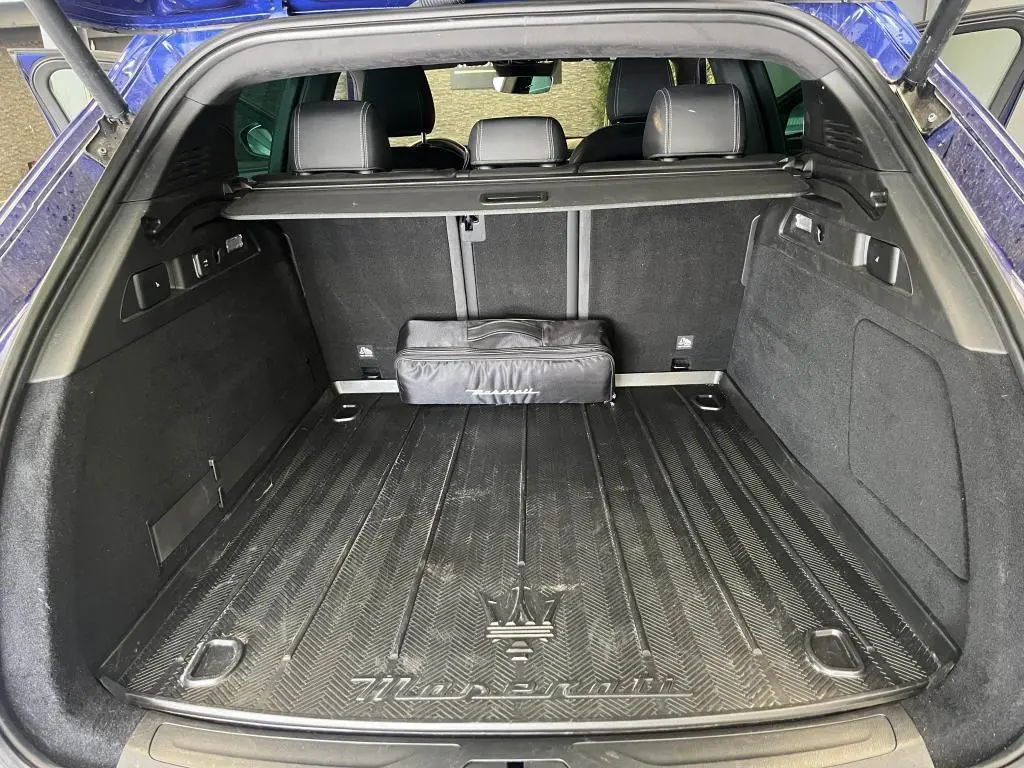 Coffre ouvert d’un Maserati Grecale bleu, vue arrière montrant tapis de coffre avec logo Trident et sac noir.