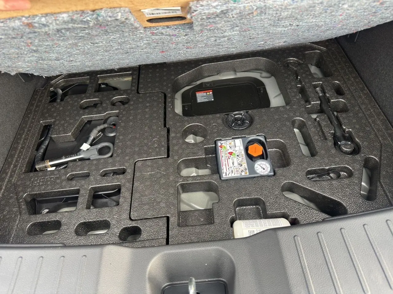 Gros plan sur le coffre ouvert d'un Nissan Juke gris squale, montrant le rangement pour outils et compresseur intégré.