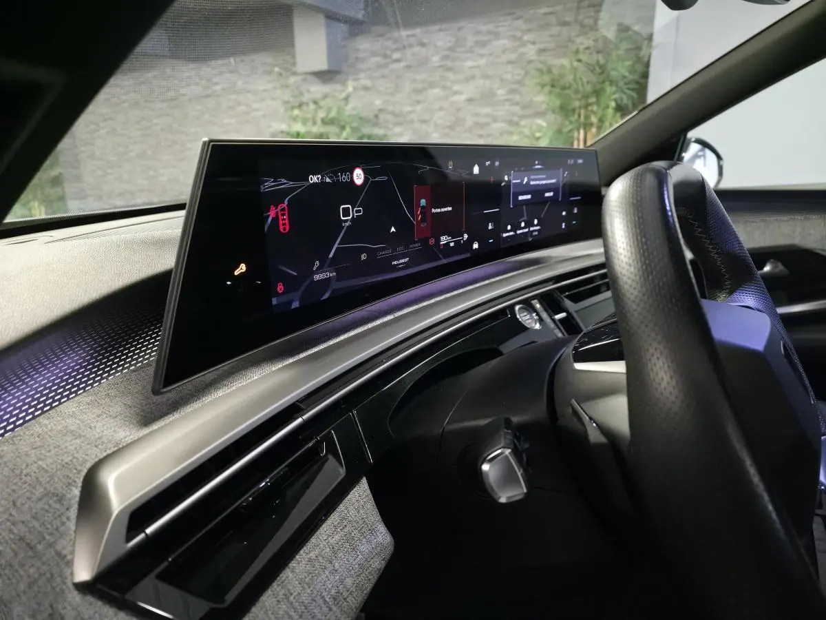 Vue intérieure côté conducteur du tableau de bord numérique panoramique et volant perforé noir du Peugeot 3008 gris 2025.