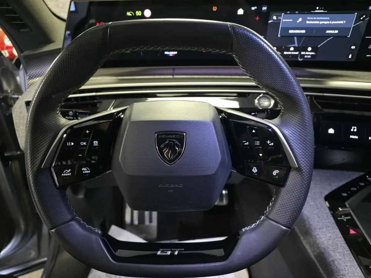 Vue rapprochée du volant cuir noir perforé avec logo Peugeot et badge GT du Peugeot 3008 gris hybride 2025.
