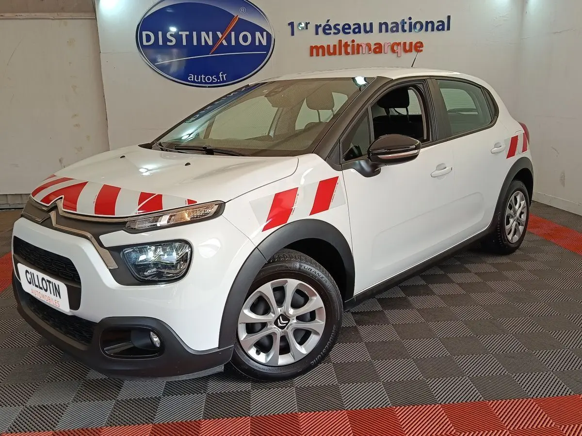 Vue 3/4 avant gauche d'une Citroën C3 blanche avec bandes rouges distinctives sur capot et ailes, en intérieur showroom.
