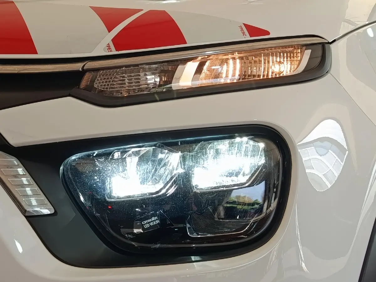 Gros plan sur le phare avant droit allumé d'une Citroën C3 blanche, mettant en valeur les LED et le clignotant orange.
