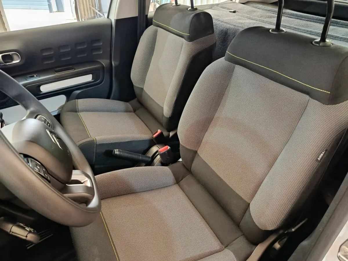 Vue intérieure côté conducteur de la Citroën C3 2021 blanche, montrant les sièges tissu gris avec surpiqûres jaunes et le volant multifonctions.