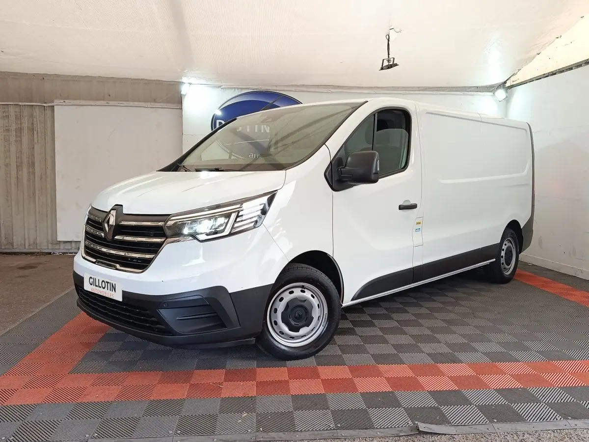 Renault Trafic Fourgon blanc vu en 3/4 avant droit, avec calandre chromée et phares LED allumés dans un showroom.