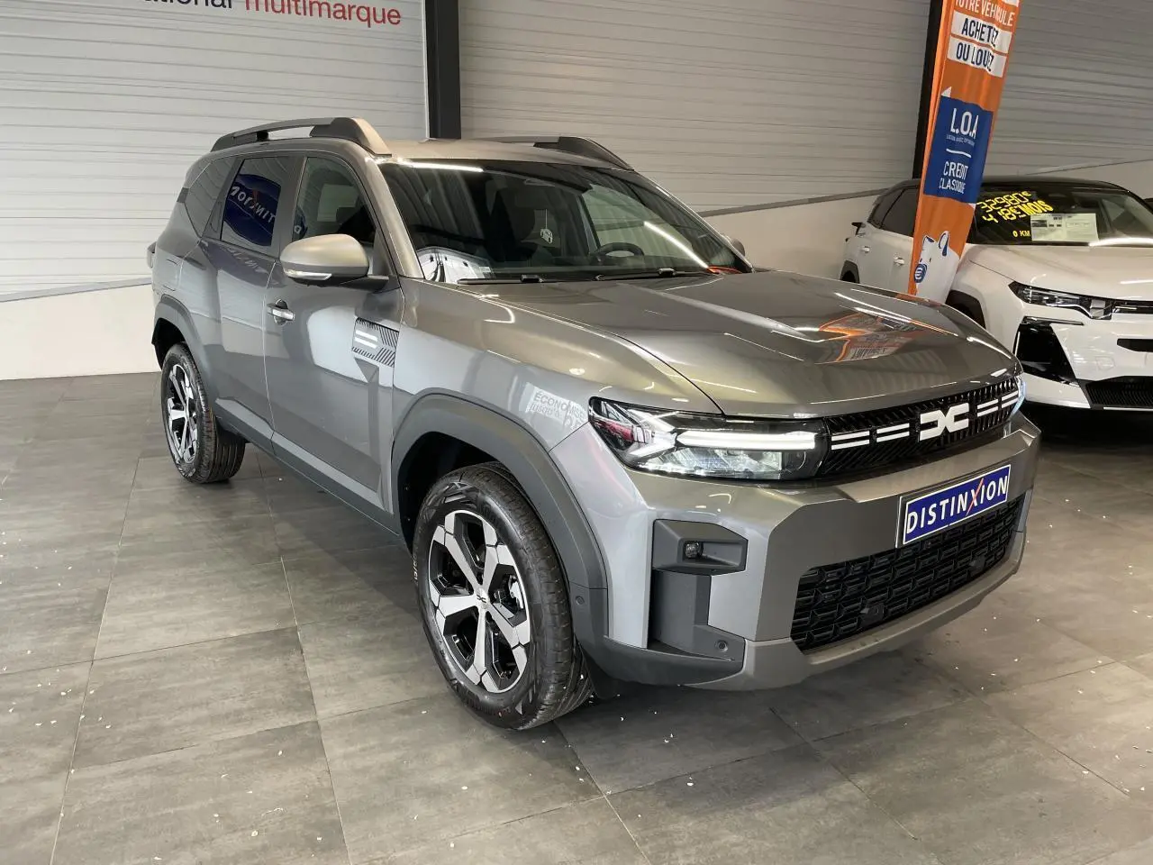 SUV Dacia Bigster gris schiste en 3/4 avant droit, avec jantes alliage diamantées et barres de toit visibles.