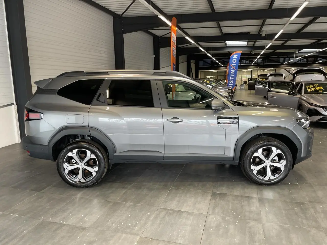 Vue de profil droit du Dacia Bigster gris schiste en showroom, avec jantes alliage 18 pouces et barres de toit longitudinales.