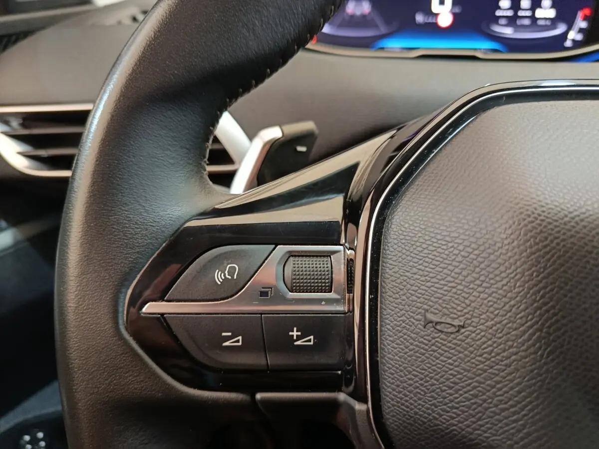 Gros plan sur les commandes vocales et volume du volant cuir noir du Peugeot 3008 2022, tableau de bord digital en arrière-plan.
