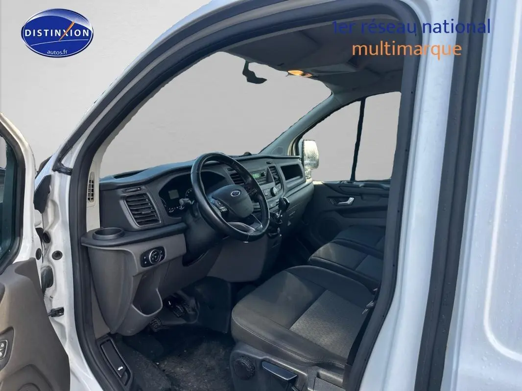 Vue intérieure côté conducteur du Ford Transit Custom blanc 2021, montrant le tableau de bord et les sièges en tissu noir.