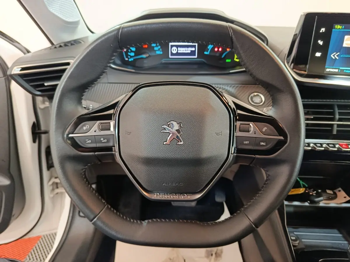 Vue rapprochée du volant cuir multifonctions d’une Peugeot 208 électrique blanche, avec tableau de bord et écran tactile en arrière-plan.