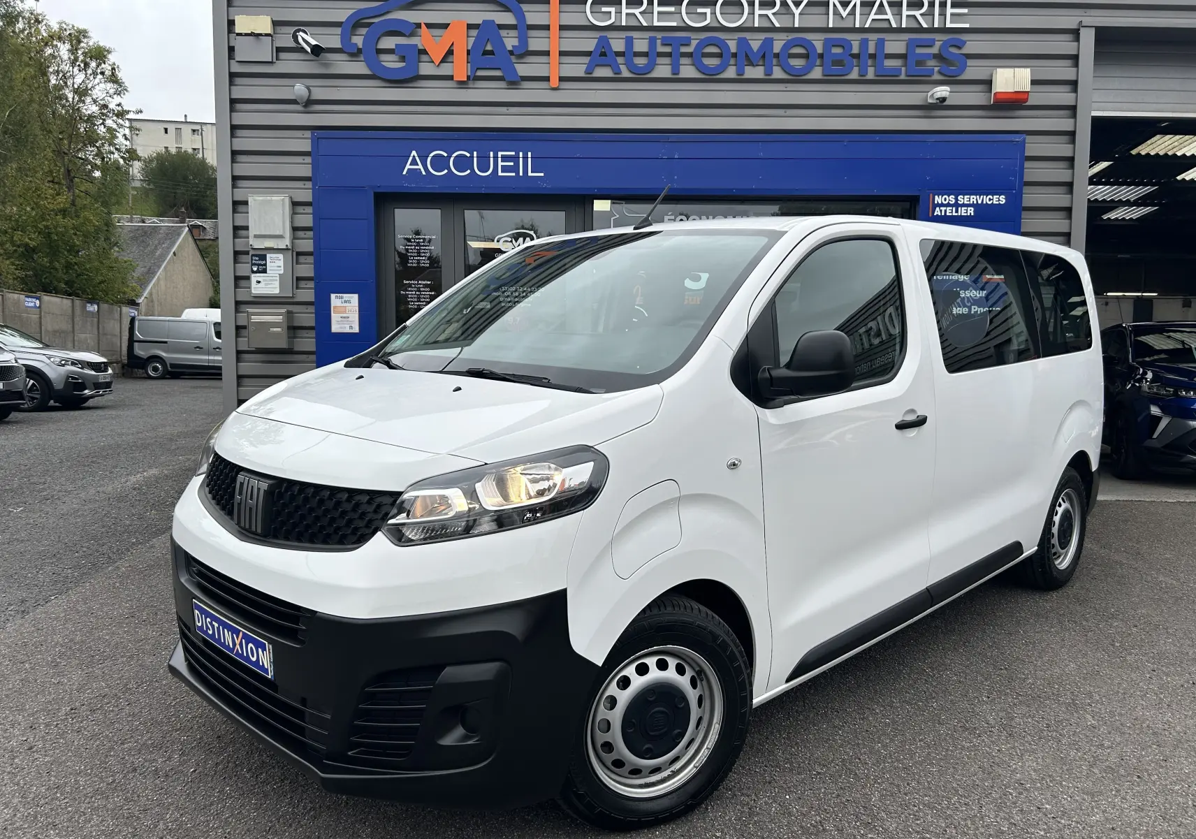 Fiat Scudo Combi blanc en 3/4 avant droit, avec calandre noire et jantes acier, devant un garage automobile.