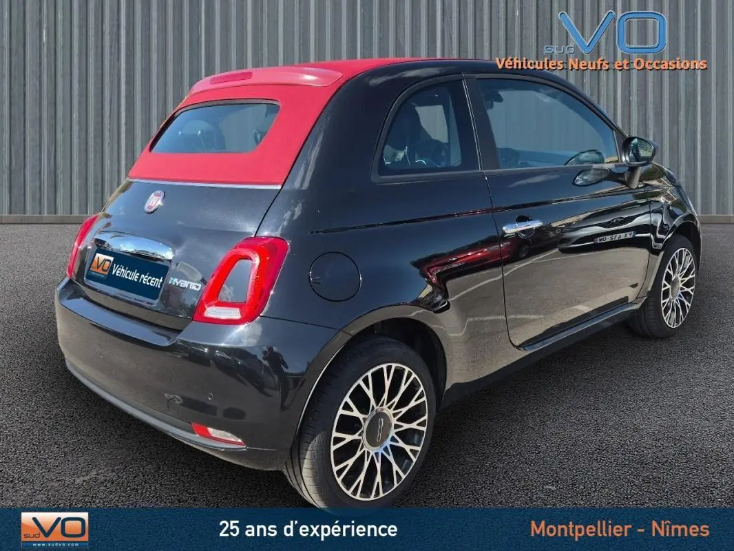 Vue 3/4 arrière droite d'une FIAT 500C noire 2024 avec toit rouge et roues alliage, version hybride essence avec roue de secours visible.