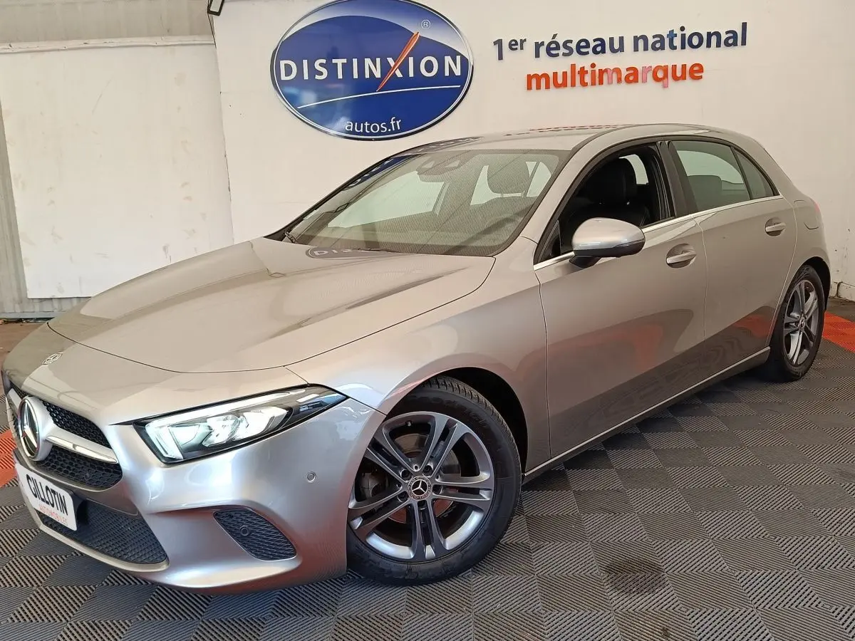 Vue 3/4 avant d'une Mercedes Classe A 180 Business Line gris clair avec jantes alliage et phares LED allumés.