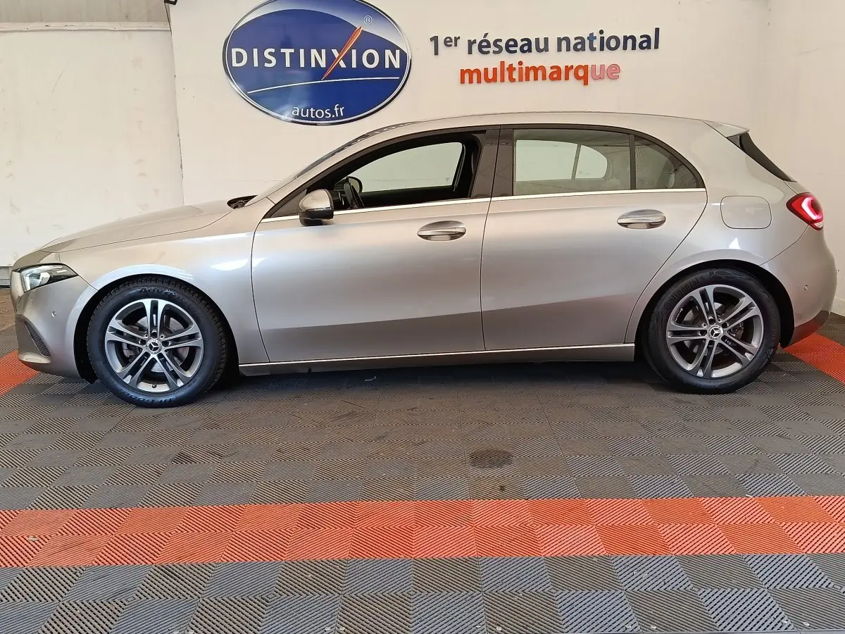 Vue de profil côté gauche d'une Mercedes Classe A 180 Business Line gris clair, avec jantes alliage et feux arrière LED allumés.